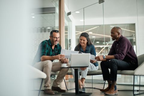 Three coworkers seated at a small table smiling and looking at papers